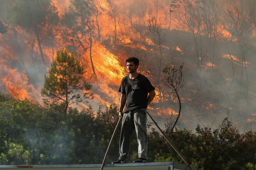 A volunteer stands on a roof as flames rise from a wildfire burning in Stamata, near Athens, Greece